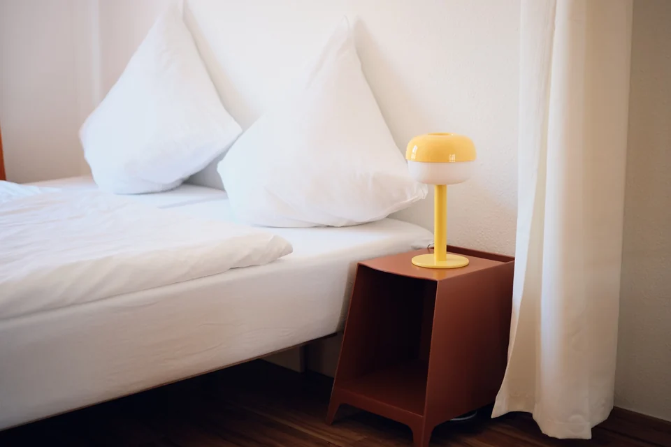 Weiß bezogenes Bett mit Kissen und einer gelben Leuchte auf dem Nachttisch im Wohnbereich. – Apartment Mittendrin.