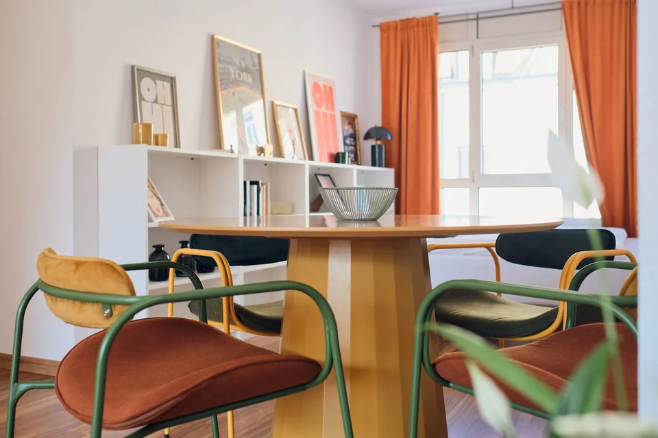 Runder Esstisch mit farbigen Stühlen vor einem Regal und einem Fenster mit orangefarbenen Vorhängen. – Apartment Mittendrin.