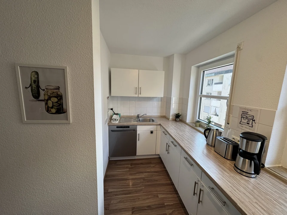 Vom Eingang aus fällt der Blick auf die helle Küche mit Holzarbeitsfläche, Fenster und Geräten. – Apartment Mittendrin.