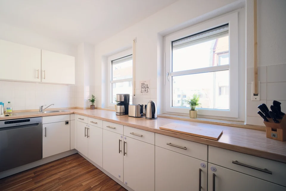 Helle Küchenfronten, Holzarbeitsfläche, Spüle und Tageslicht durch das große Fenster. – Apartment Mittendrin.