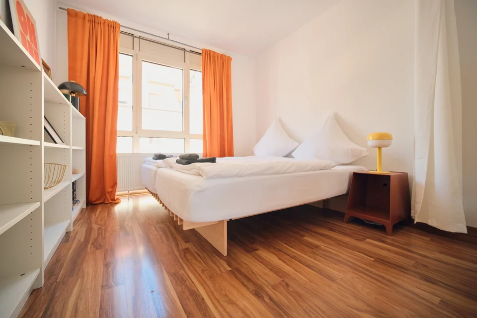 Ein Doppelbett im Wohnbereich, neben Regal, Nachttisch und großem Fenster. – Apartment Mittendrin.
