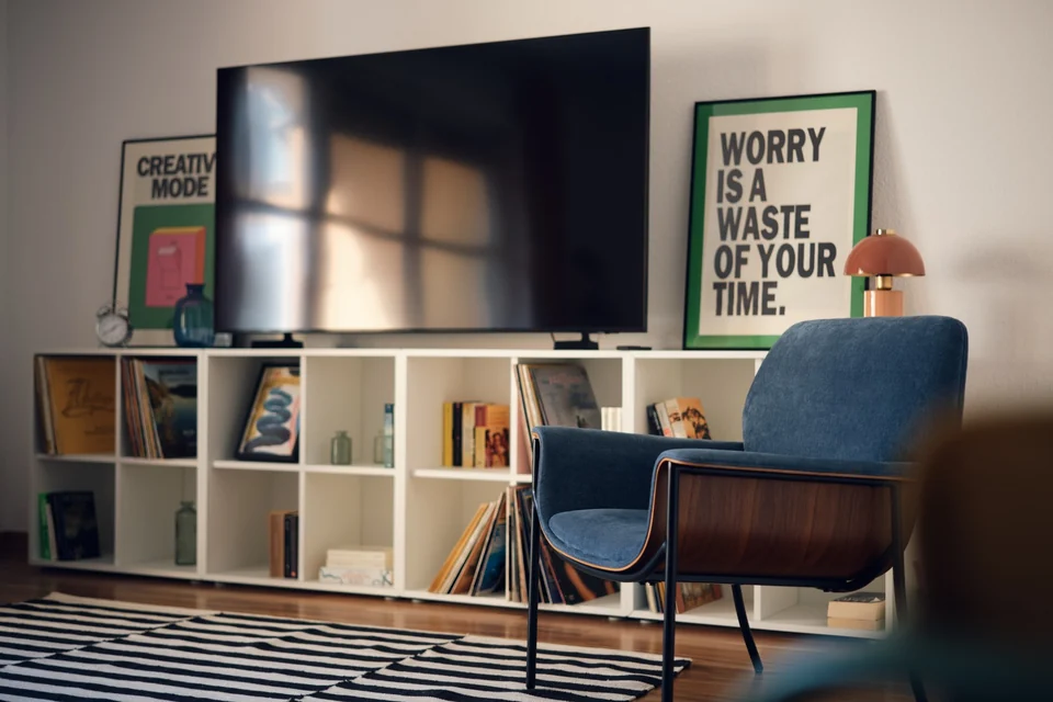 Großer Fernseher über einem offenen Regal und ein blauer Sessel im Wohnbereich. – Apartment Mittendrin.
